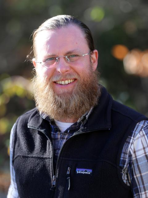 Nathan Thomas is a man with long hair pulled back, a long reddish blond beard, who is wearing glasses, a blue plaid flannel with a fleece vest over it